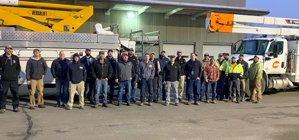 A group of WPS and We Energies employees standing in front of large utility vehicles early in the morning.