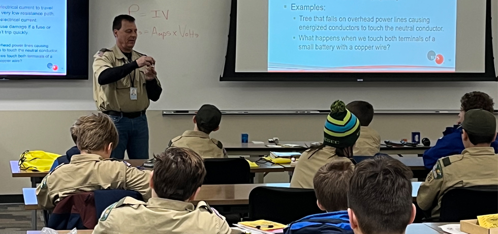 A male WPS employee and Scout leader discusses electrical components with a large group of Scouts.