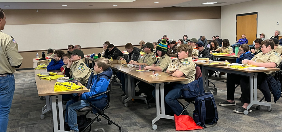A large group of Boy Scouts sit at tables in a large conference room.