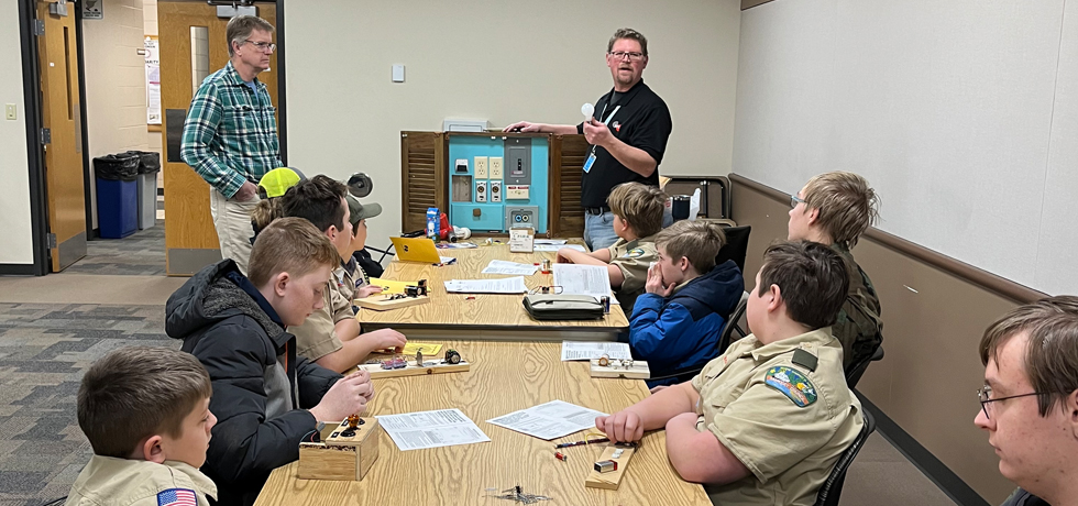 Two male WPS employees operate an electric circuit display for a small group of Boy Scouts.