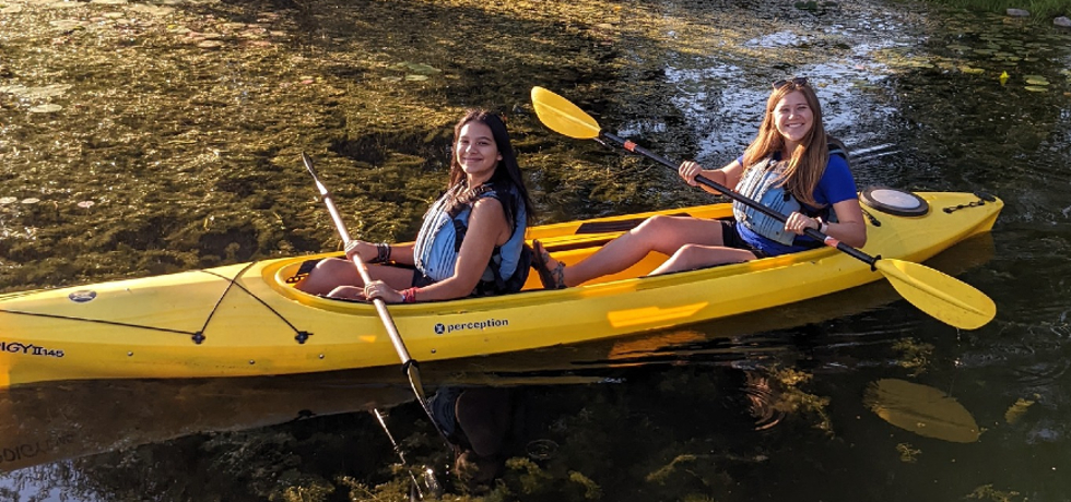 A teenage girl and a woman smiling and paddling a yellow kayak in a stream.