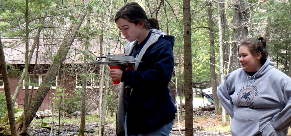 A young woman with a black jacket on stands in a wooded area looking at a monitoring device. Another young woman stands behind her.