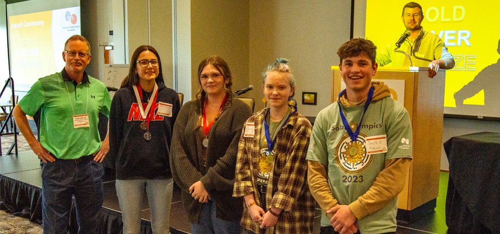 A male Solar Olympics hosts stands next to high school students who stand for a photo with the medals they won at the 27th WPS Solar Olympics.