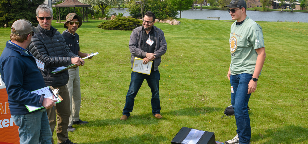 A group of adult male judges speaks with a male student about a solar cooker placed on a grass lawn.