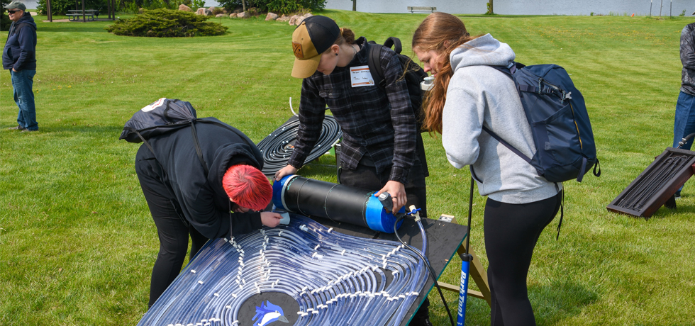 Three female high school students set up a solar water heater device on a grass lawn at the 27th annual WPS Solar Olympics