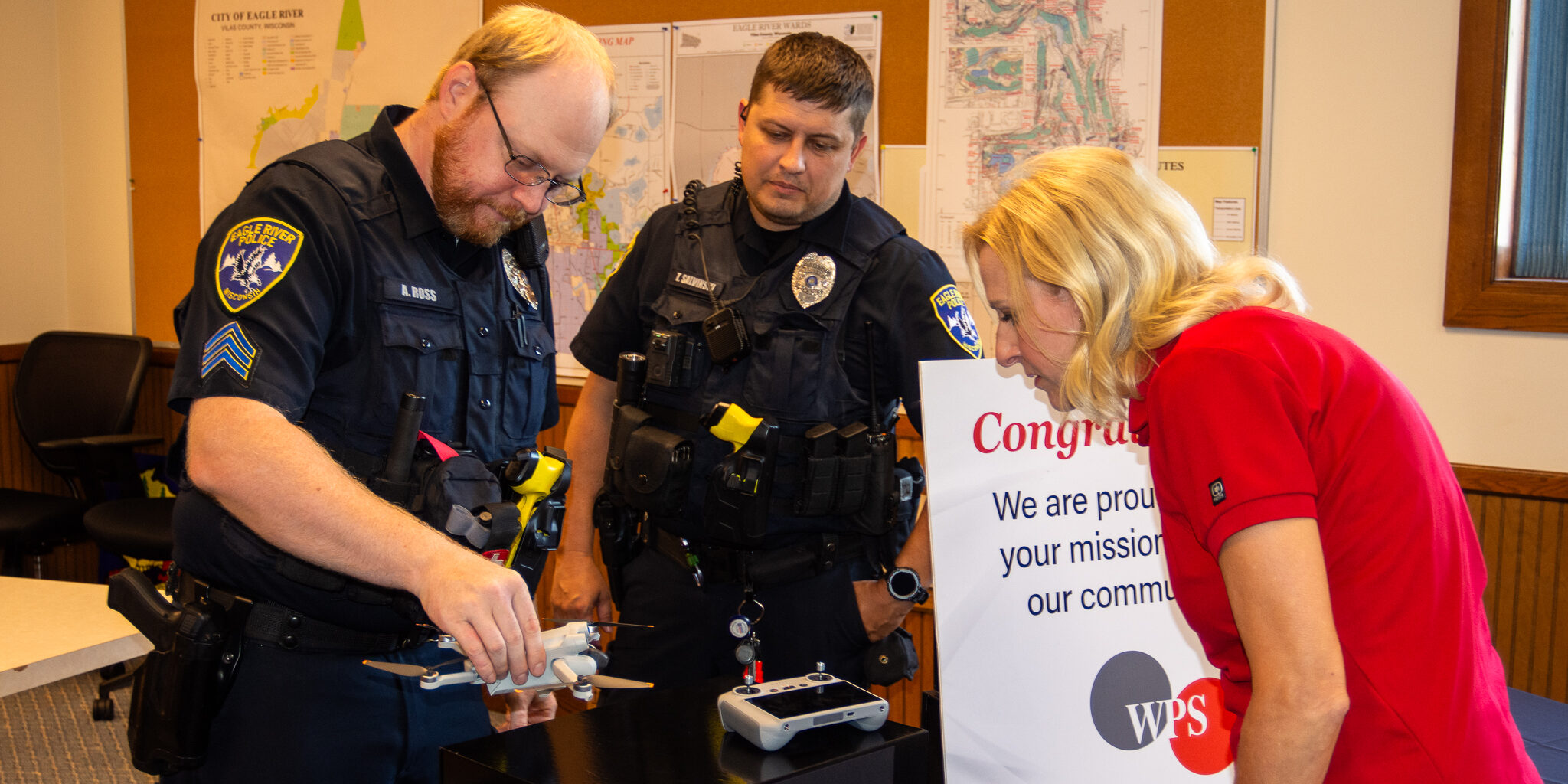 A pair of police officers and a woman from the WPS Foundation look at a small aerial drone inside a large room.