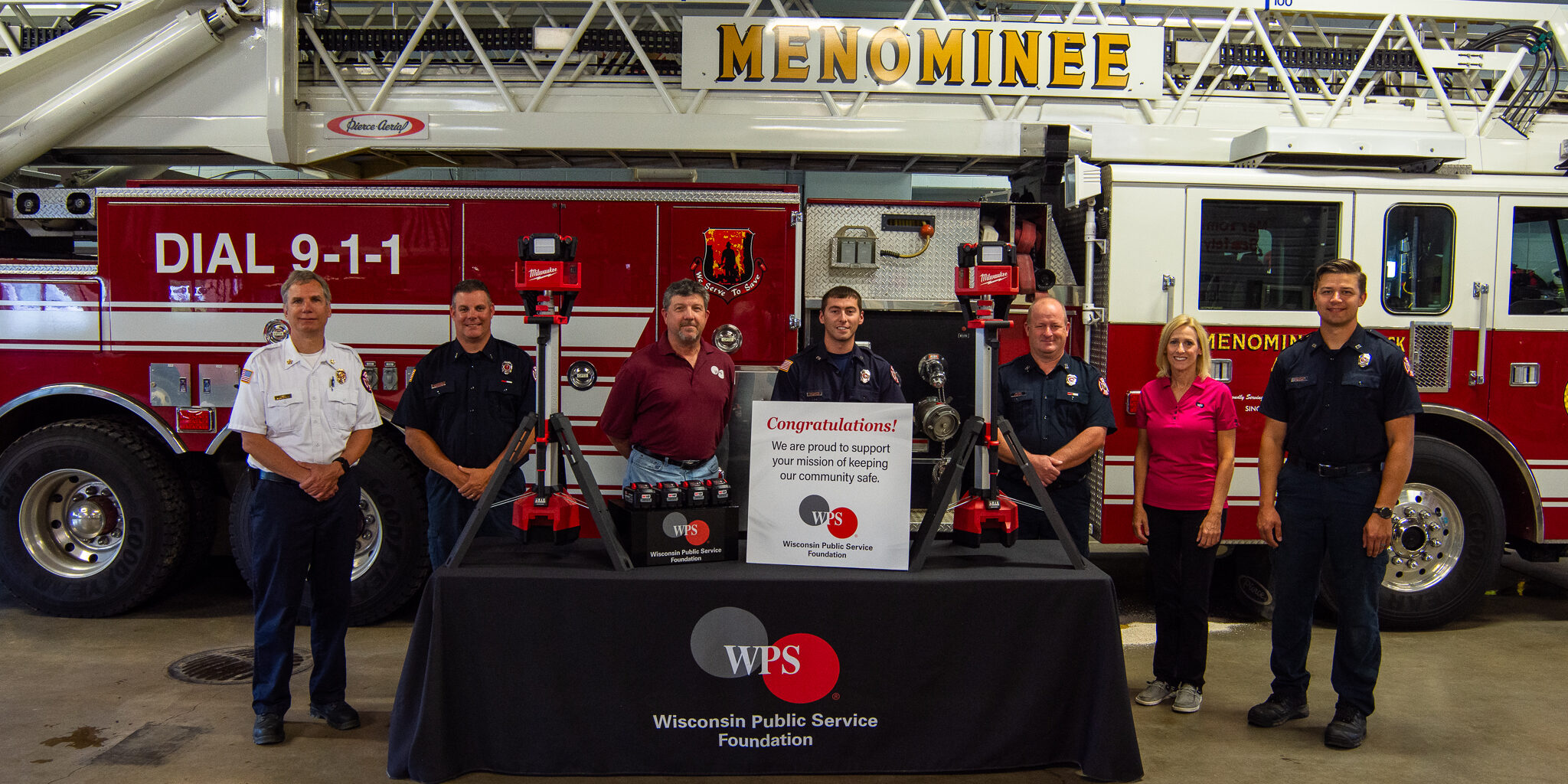 Firefighters and a woman stand behind a table with a WPS Foundation logo on it and new emergency scene lighting equipment resting on the table.
