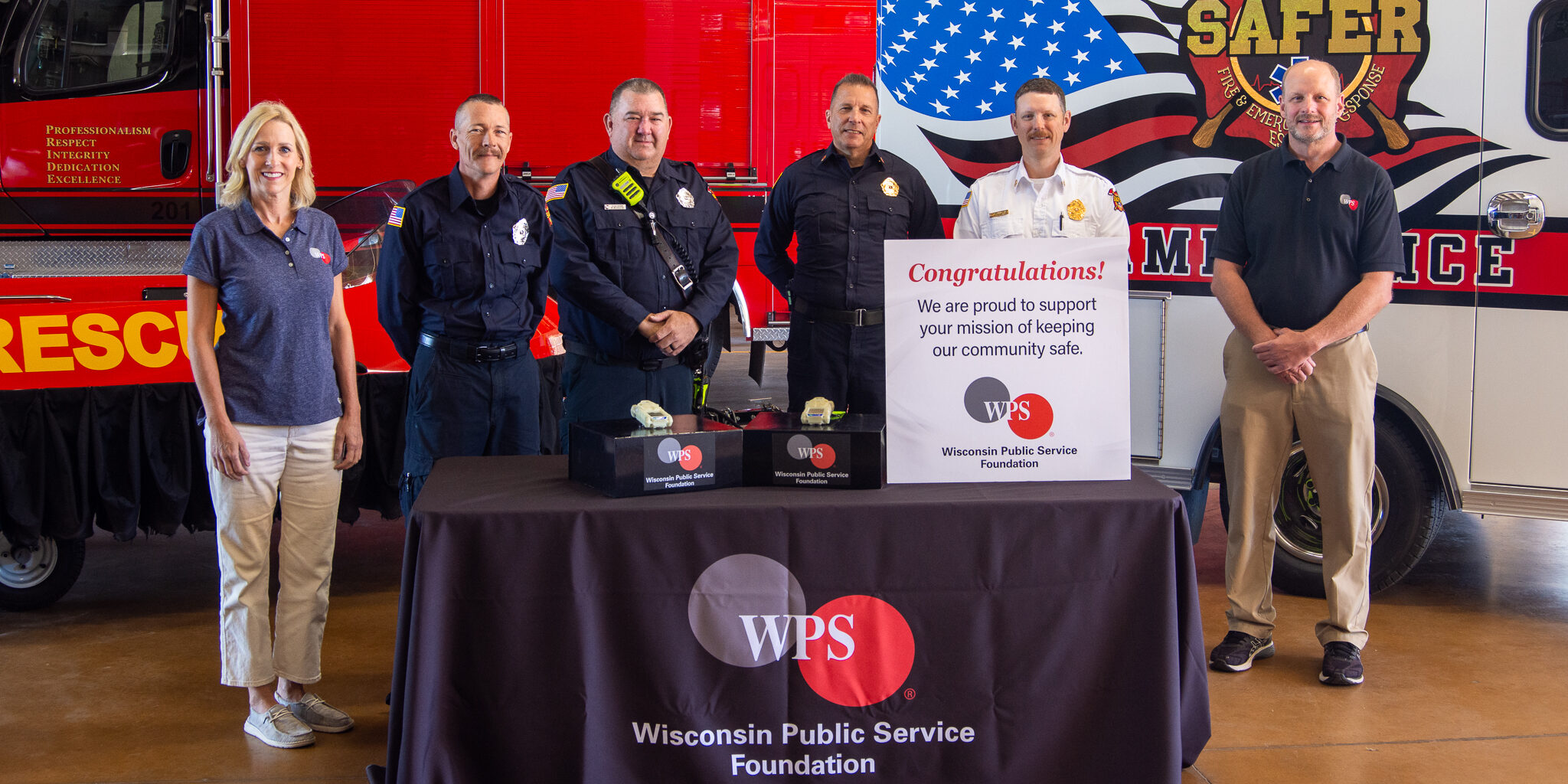 Firefighters and a man and woman from the WPS Foundation stand around a table with a sign and small multi-gas detectors sitting on it.