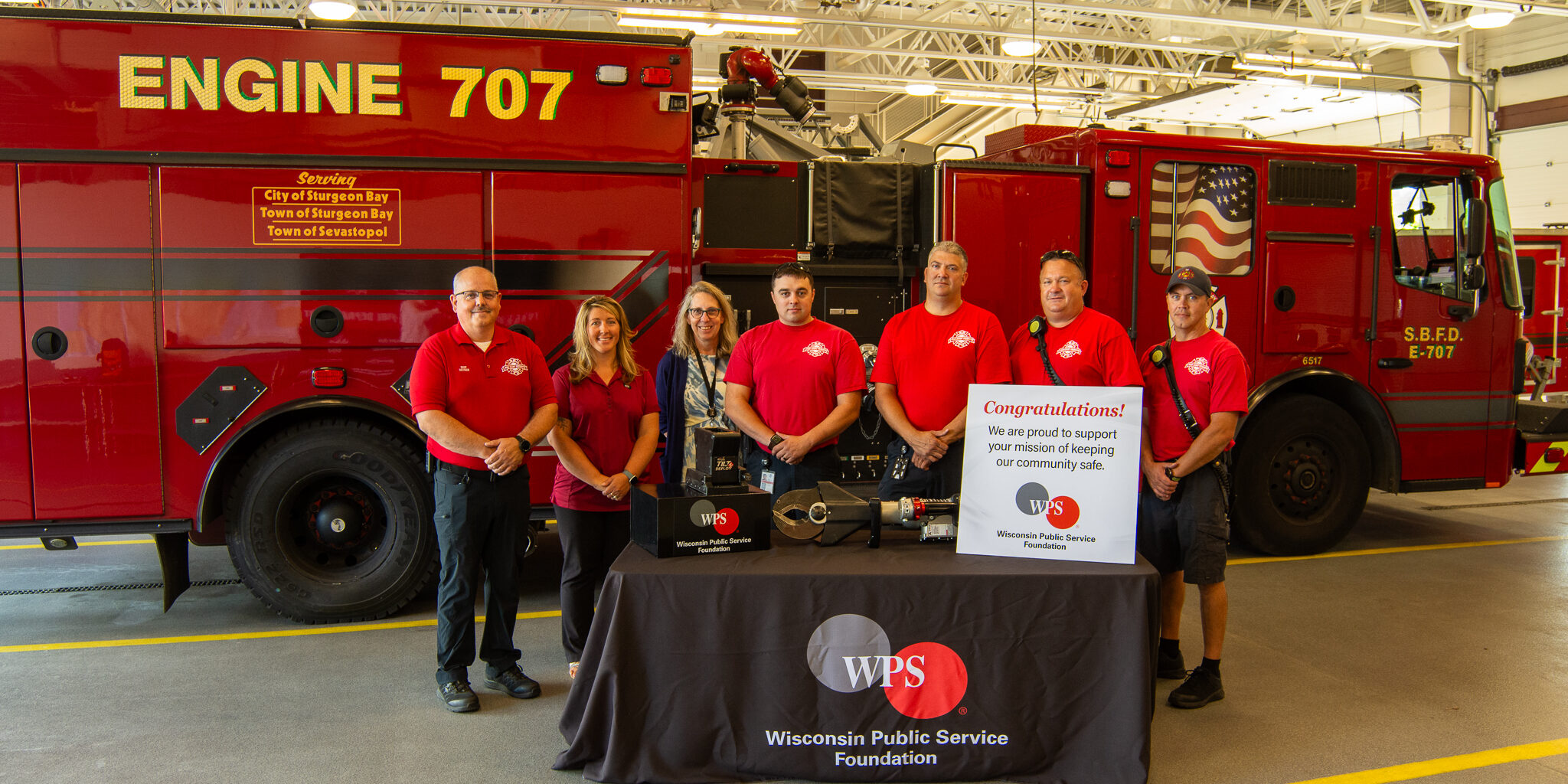 Firefighters and a woman stand behind a table with a WPS Foundation logo on it and a new heavy-duty equipment mount resting on it.