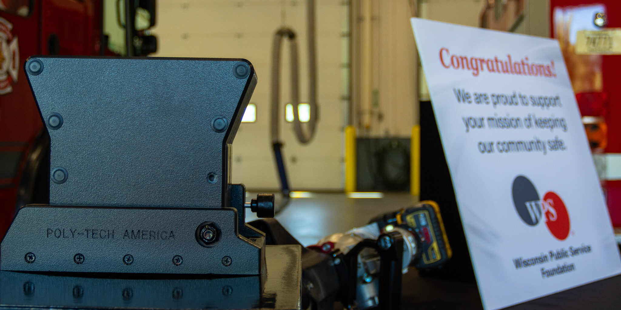 A black heavy-duty tool mount sits on a table in front of a congratulatory sign inside a fire department garage.
