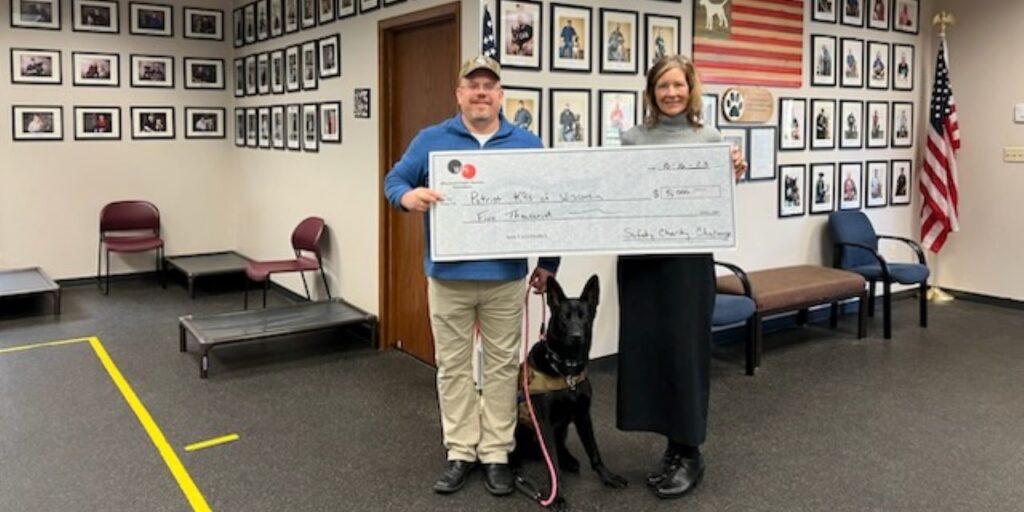 A man and woman hold a ceremonial donation check with a black dog in between them.
