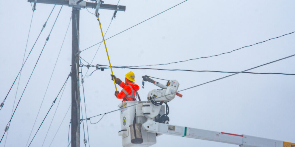 A lineworker in an orange coat works on power lines on a cloudy day.