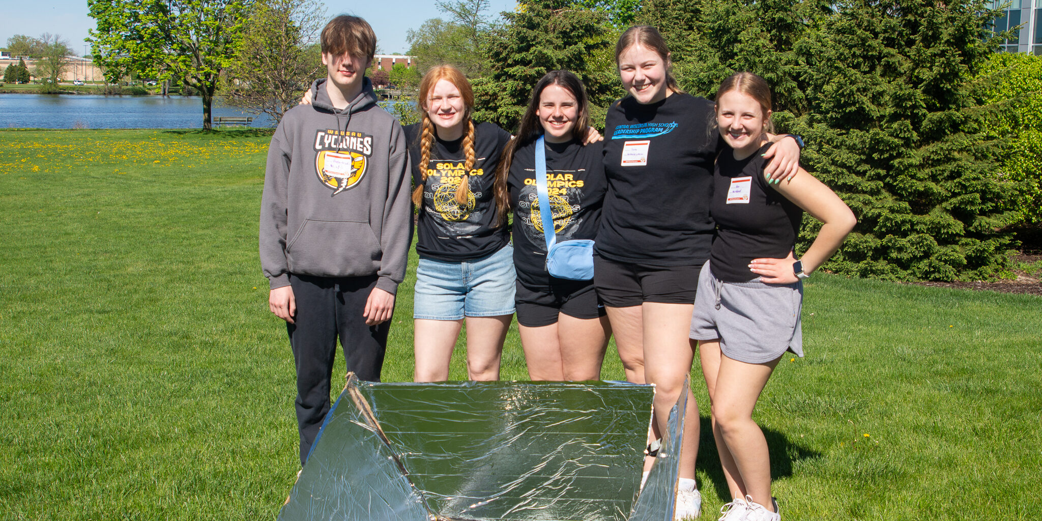 A group of high school students takes a photo behind a solar-powered cooker on a sunny day.