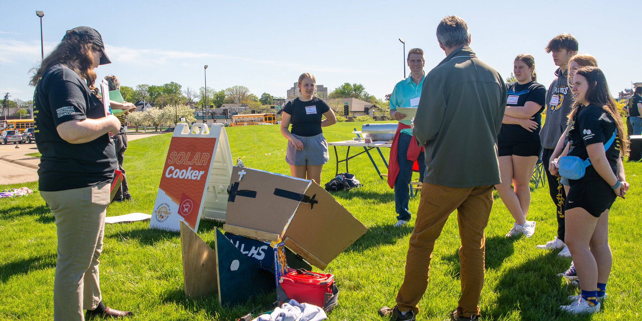 A group of high school students speaks with adults about a solar-powered food cooker on a sunny day.