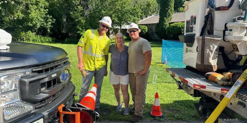 Two Wisconsin Public Service male lineworkers take a photo with a woman in a grassy area near work vehicles.