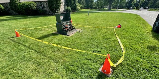 Damaged electric equipment in a grass field surrounded by yellow caution tape and orange cones.