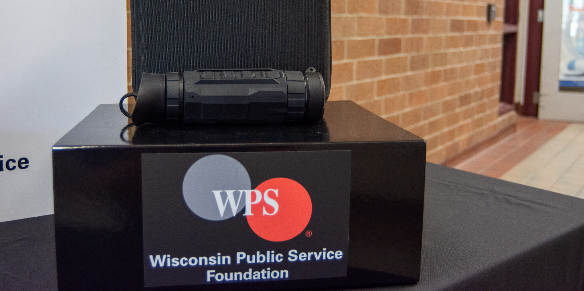 A small thermal imaging device shaped like a monocular that is lying on top of a black box with the WPS Foundation logo on it.