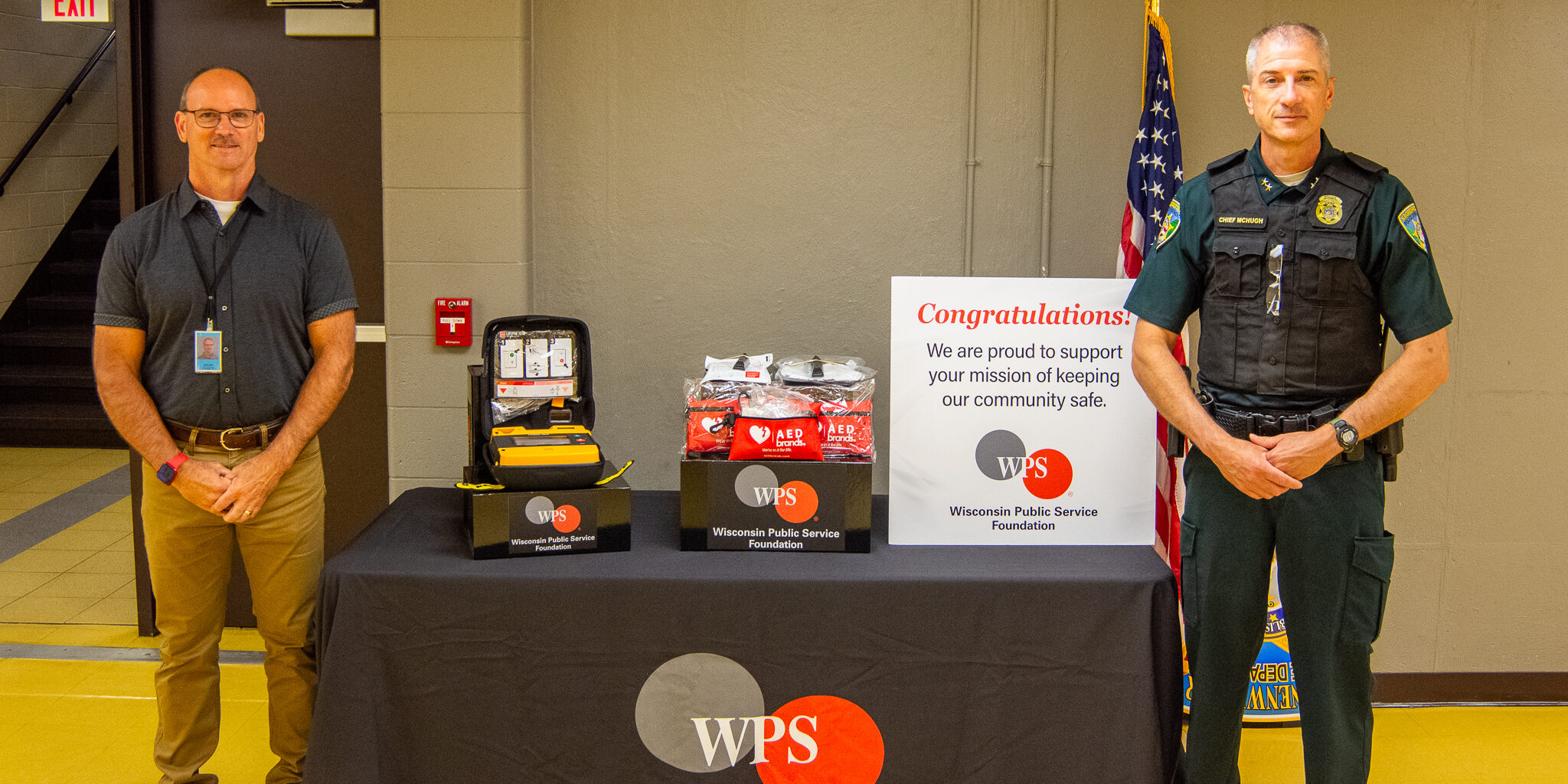 A WPS employee and the Kronenwetter Police Department chief stand on each side of a table holding an AED, AED batteries, AED pads and a congratulatory sign.