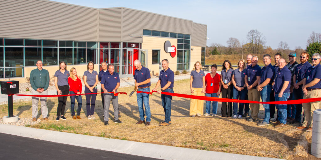 A group of WPS employees gathers behind a group of three men cutting a ceremonial red ribbon outside a building on a sunny day.