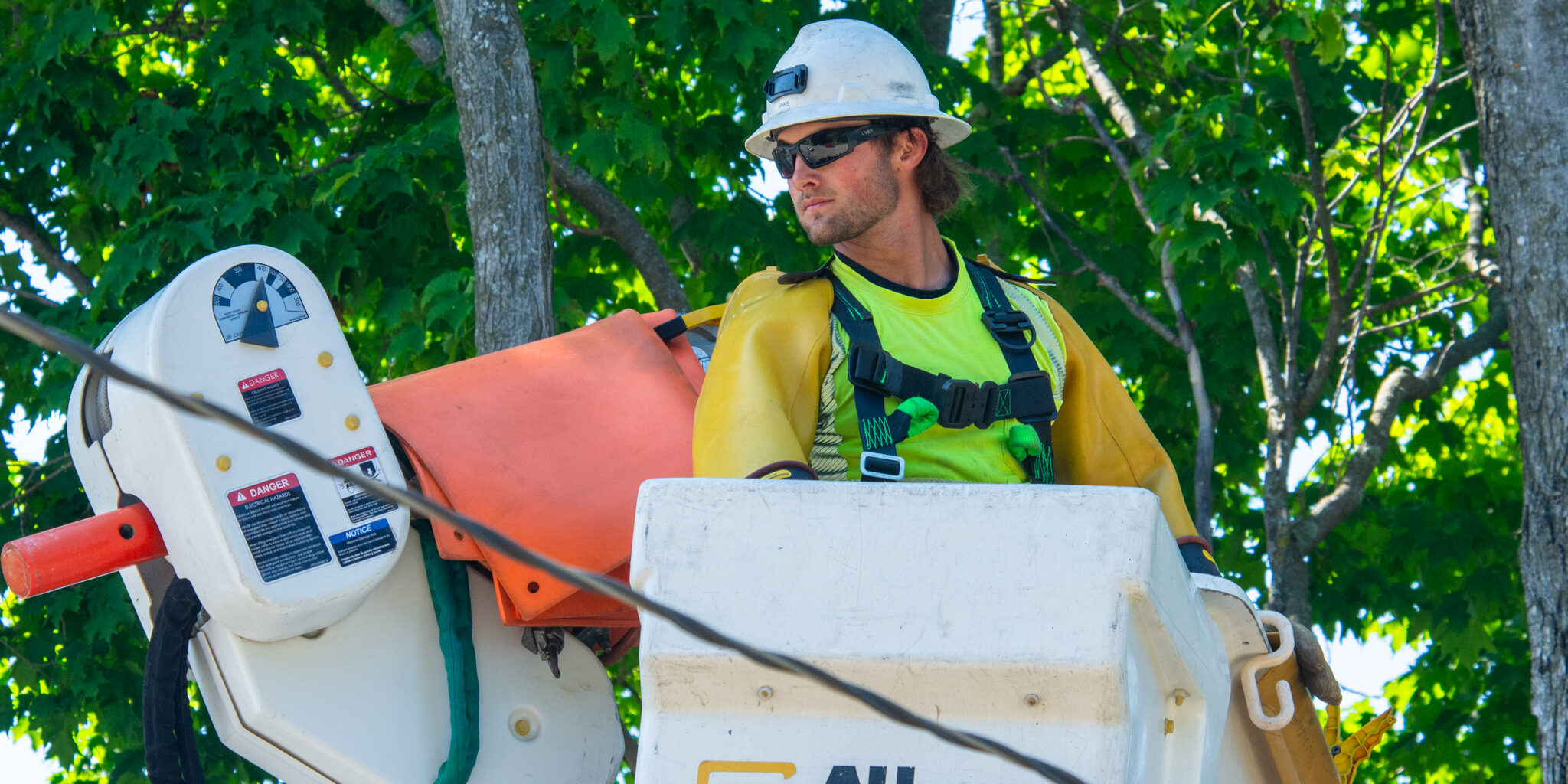 A lineworker in a utility bucket surrounded by trees looks at a power line.