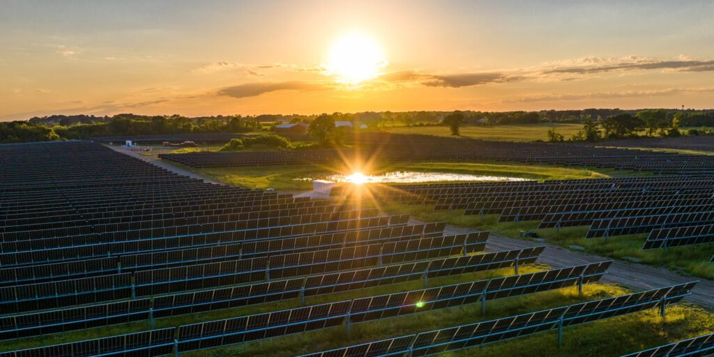 The sun sets above a large solar park surrounded by green fields.