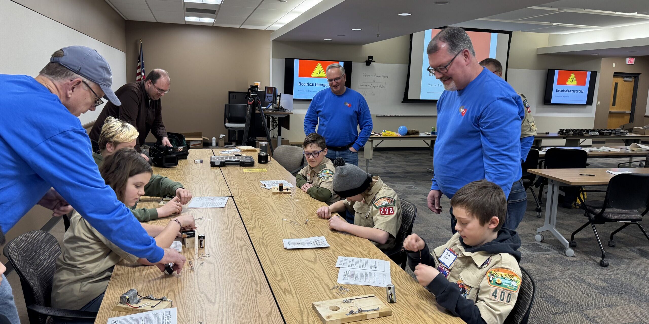 WPS employees look over electric circuits created by Scouts sitting at a long table.