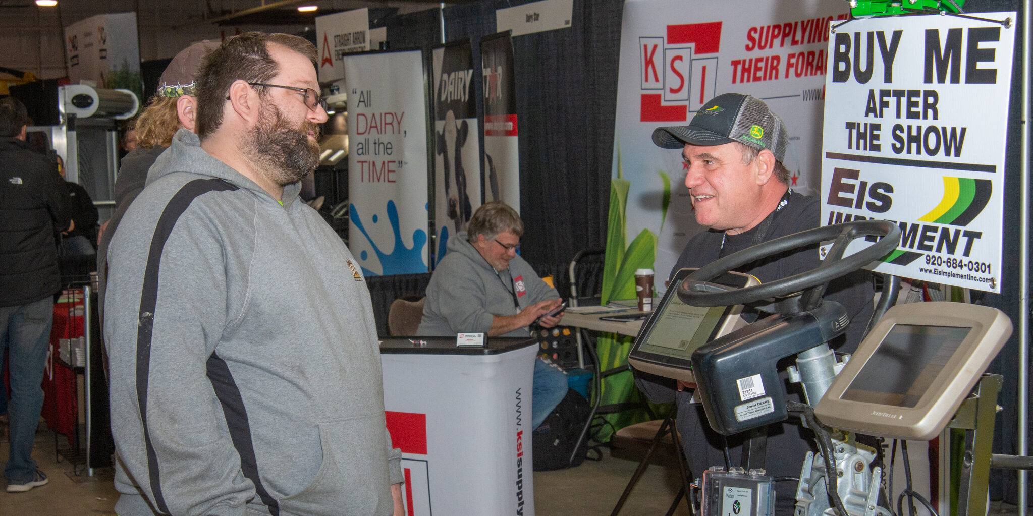 Two men talk inside an equipment booth for precision planting equipment inside a large exhibition hall.