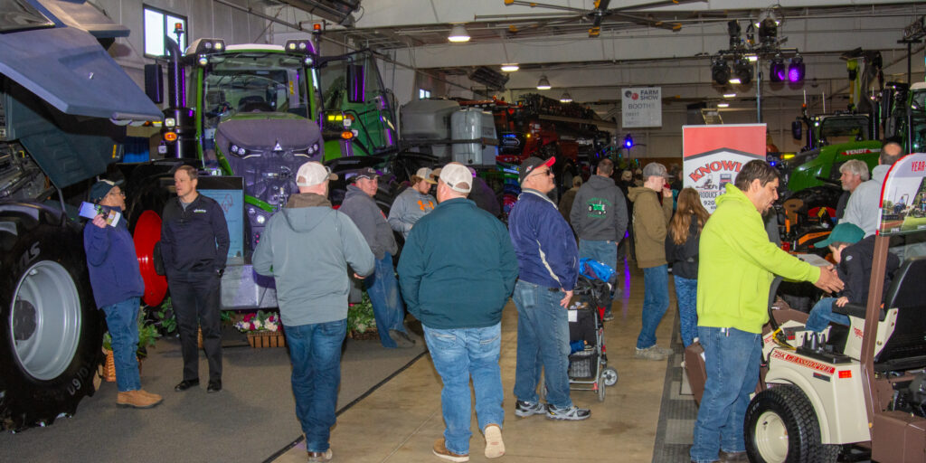 Several people walk by large farm machinery inside a large exhibition hall.