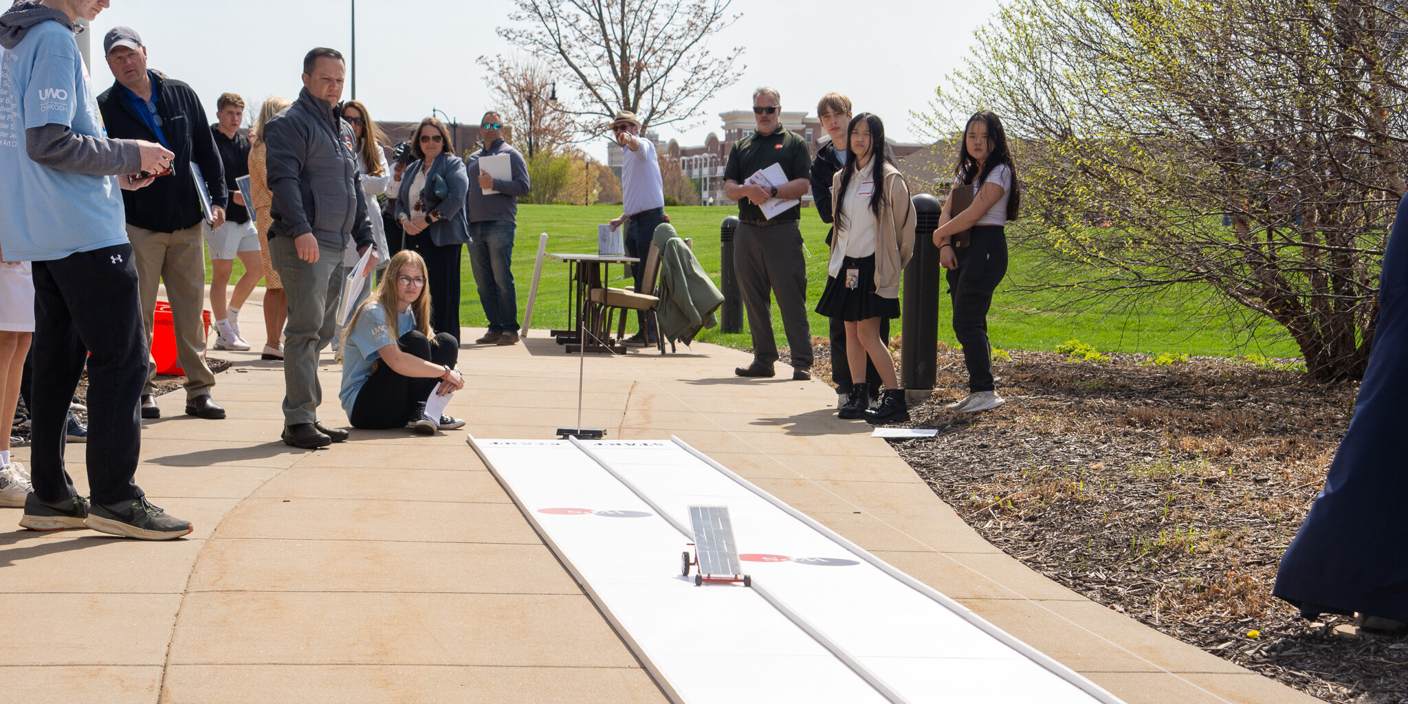 A solar-powered race car begins moving down a short track as students and judges look on.