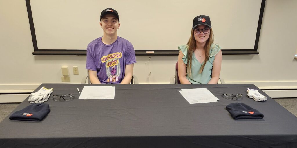 A pair of high school students smile for a photo while sitting behind a table covered in a black WPS tablecloth.