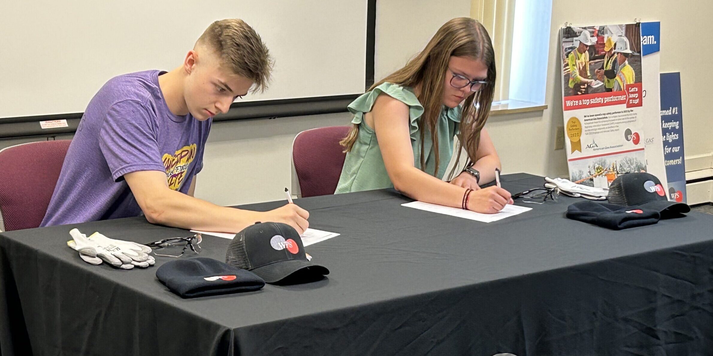 A pair of high school students sign pieces of paper while sitting at a table with WPS hats laying on it.