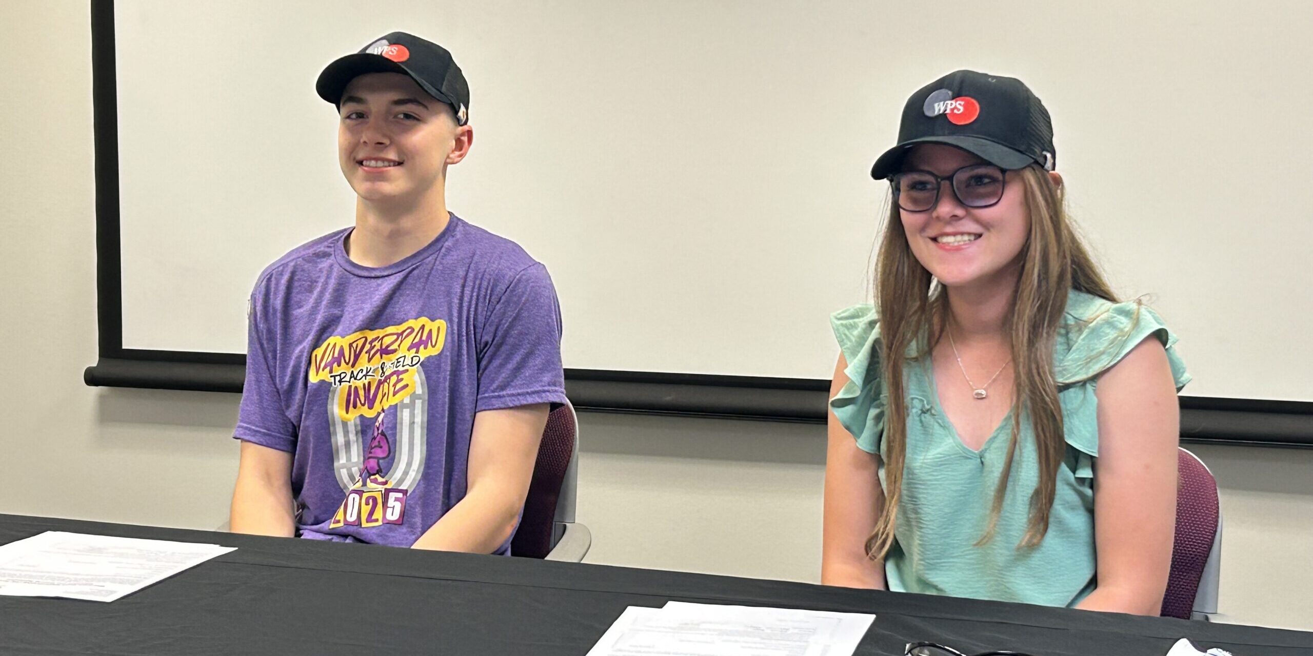 A pair of high school students smile for a photo while sitting behind a table with paperwork on it.