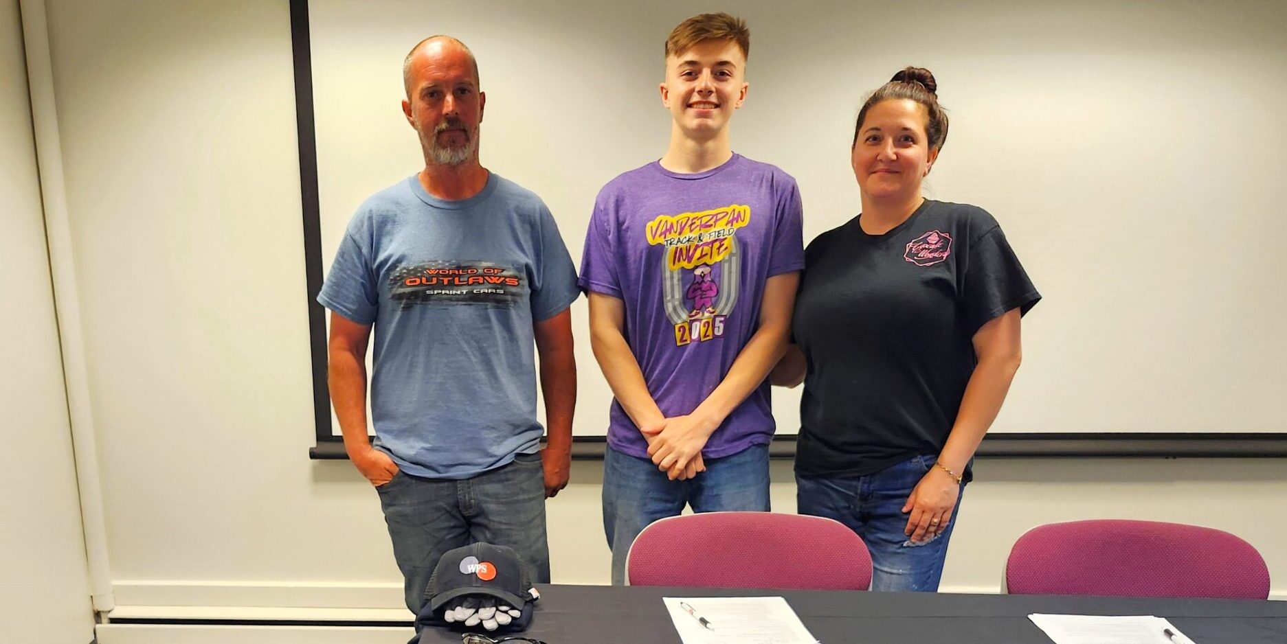 A high school student stands next to a man and woman for a photo behind a table with papers on it.