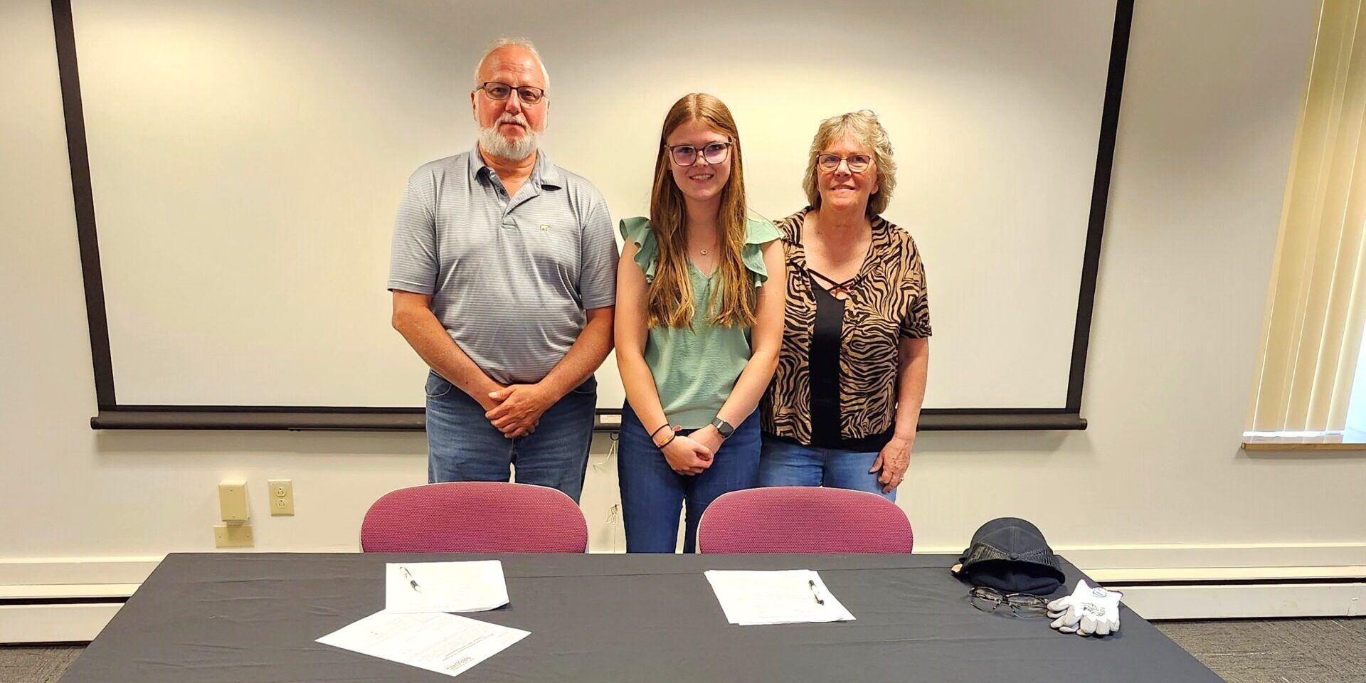 A high school student stands next to a man and woman in front of a table with papers on it.