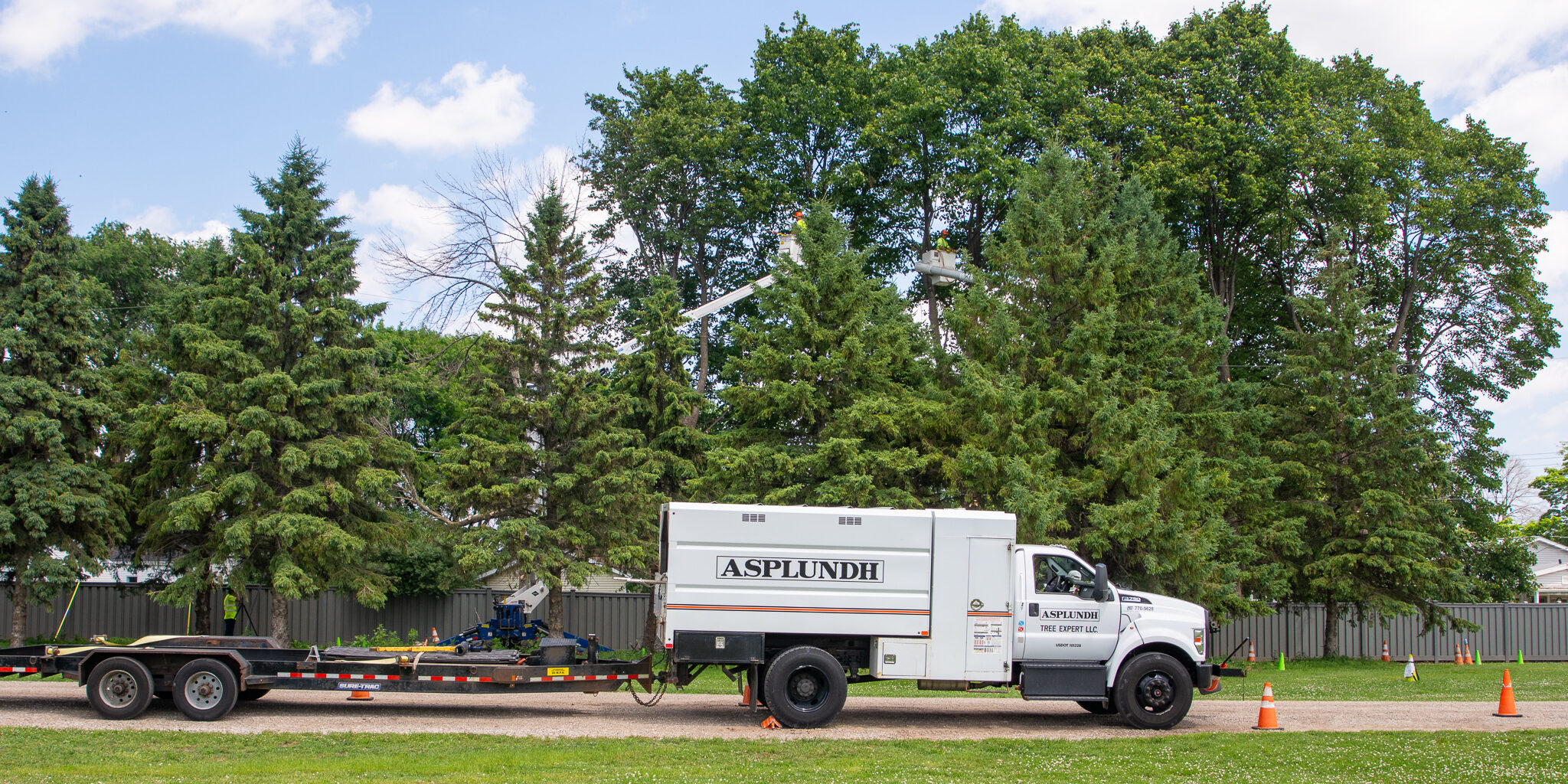 A white box truck towing a flatbed trailer parked on a gravel road. Forestry workers trim large trees in the background.