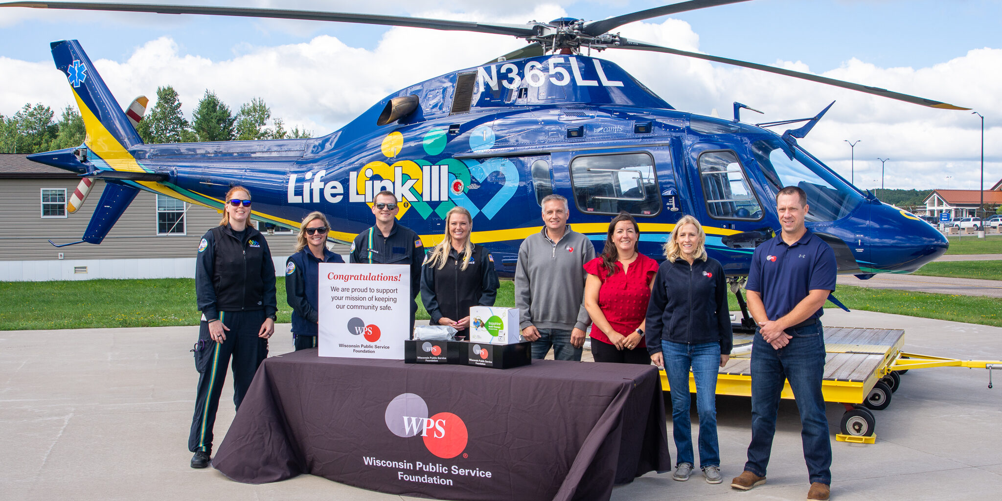 WPS employees and Life Link III staff members take a photo with a new medical infusion pump in front of an air medical transport helicopter at an airport.