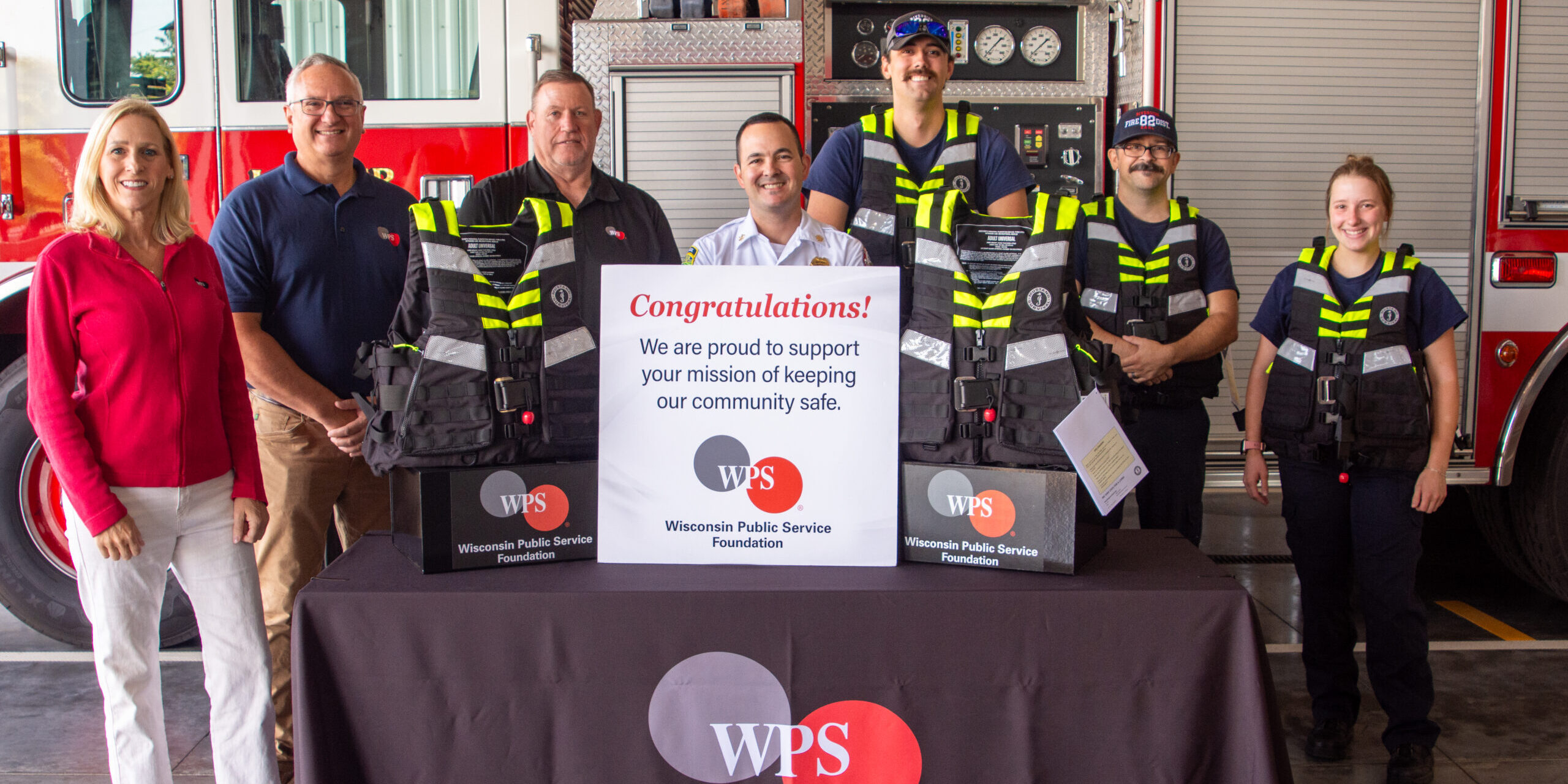 WPS employees and Riverside Fire District firefighters take a photo in front of a table covered by a WPS Foundation tablecloth and with water rescue life jackets resting on top of it.
