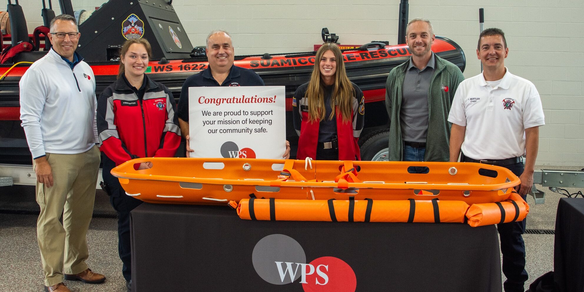 A small group of men and women stand behind an orange rescue stretcher on top of a table.