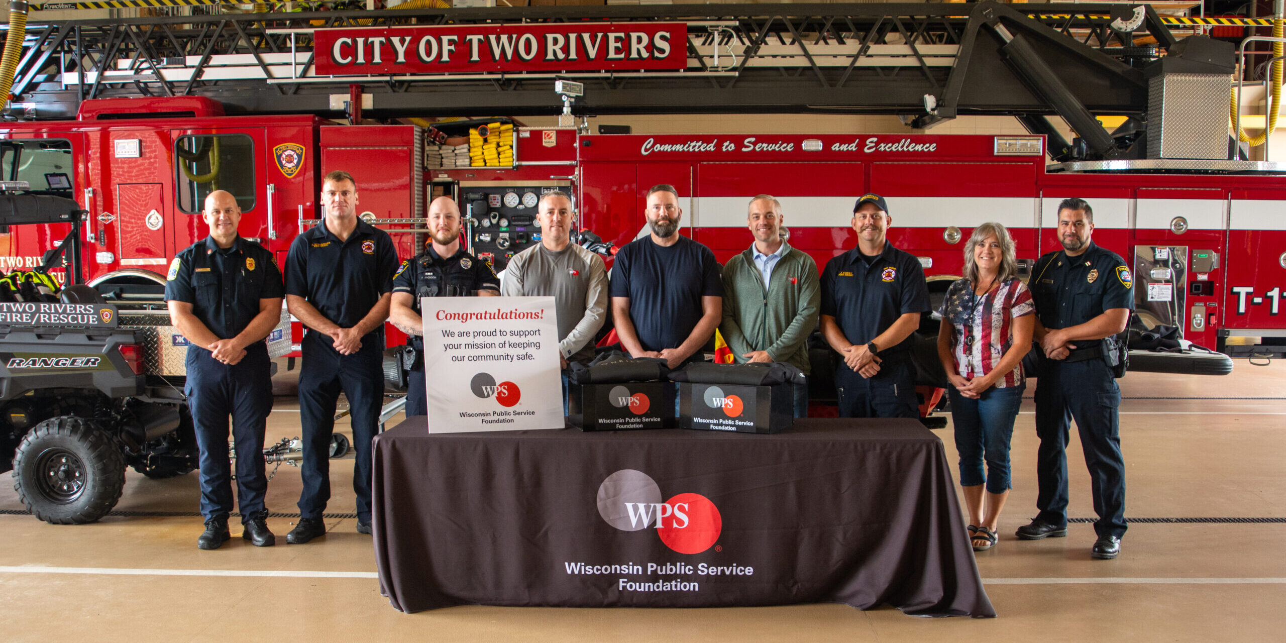 A small group of firefighters, police officers and WPS employees take a photo near a fire engine. The group is behind water rescue wetsuits lying on a table covered by a WPS Foundation tablecloth.