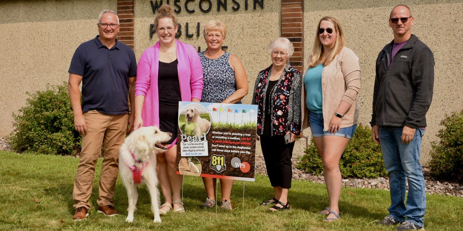 A group of people and a dog with white hair stand next to a small yard sign featuring the dog's picture, the 811 logo and the WPS logo.