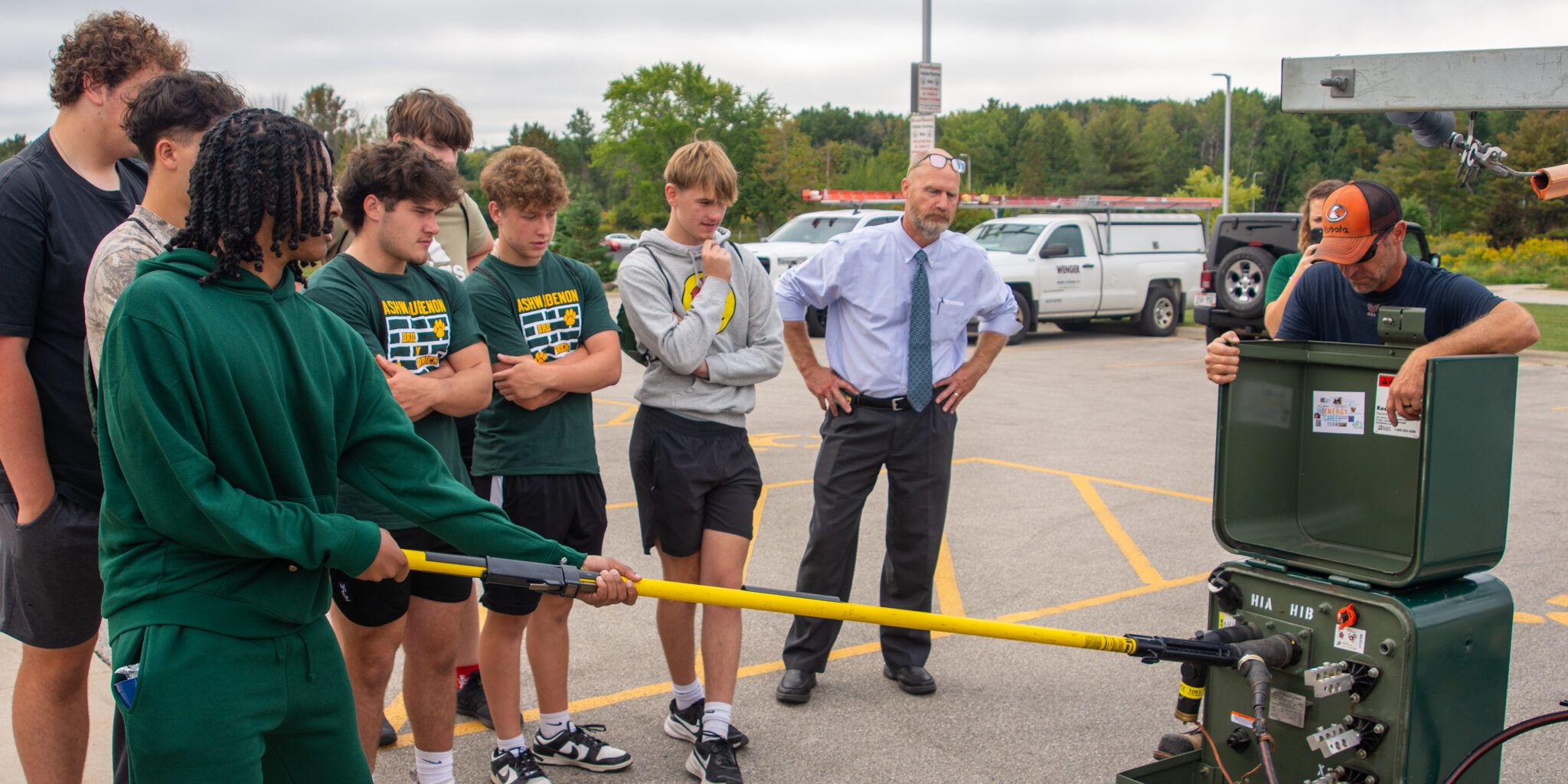 A high school student in a green sweatshirt uses a long yellow stick to pull off an electric elbow inside a mock transformer.