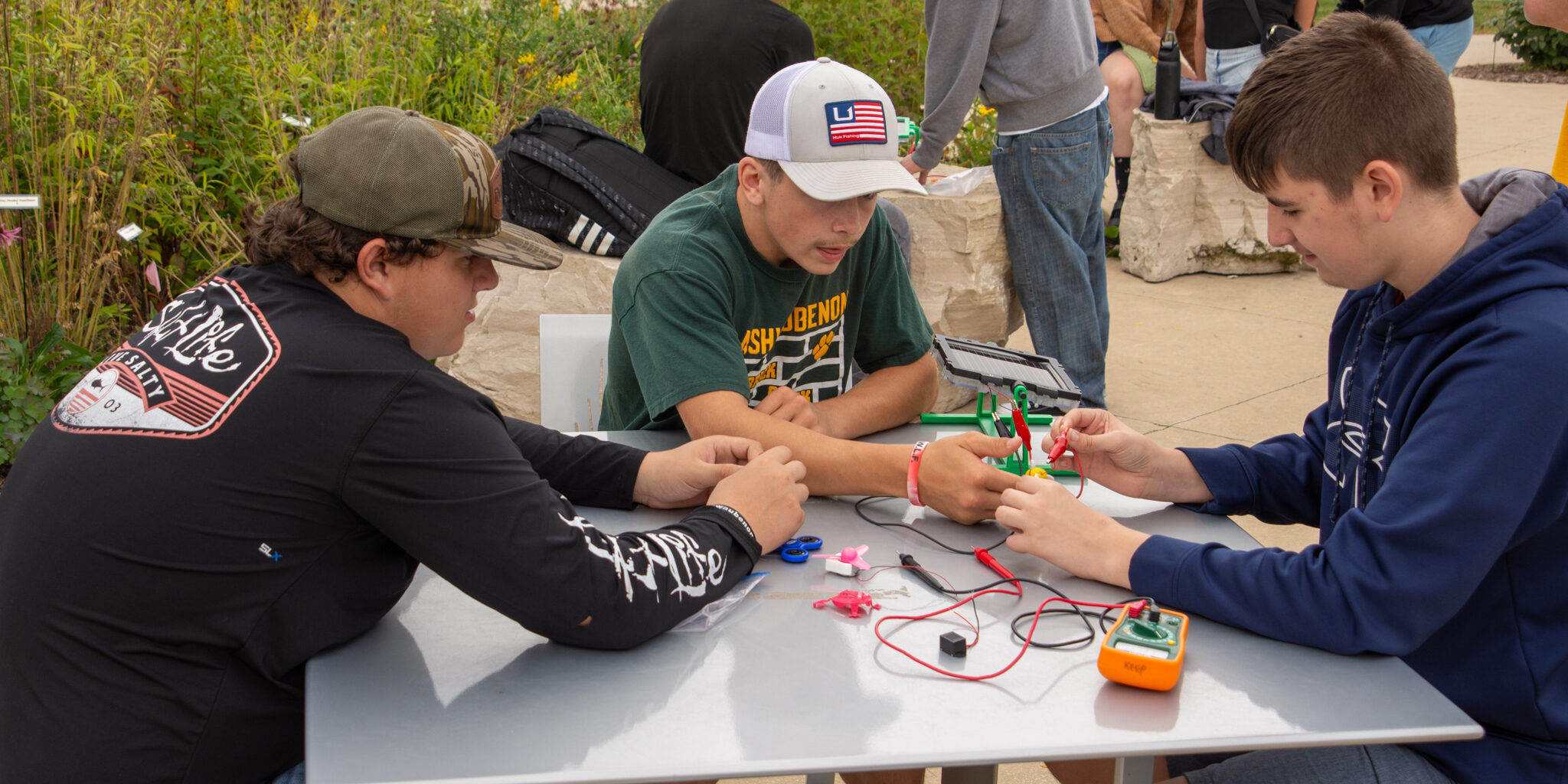 A group of three high school students connects wires to a small solar panel.