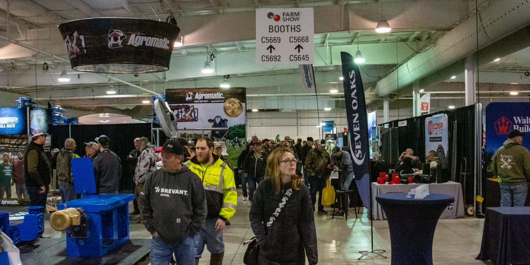 A growd walks past several exhibitors booths inside a warehouse-style building.