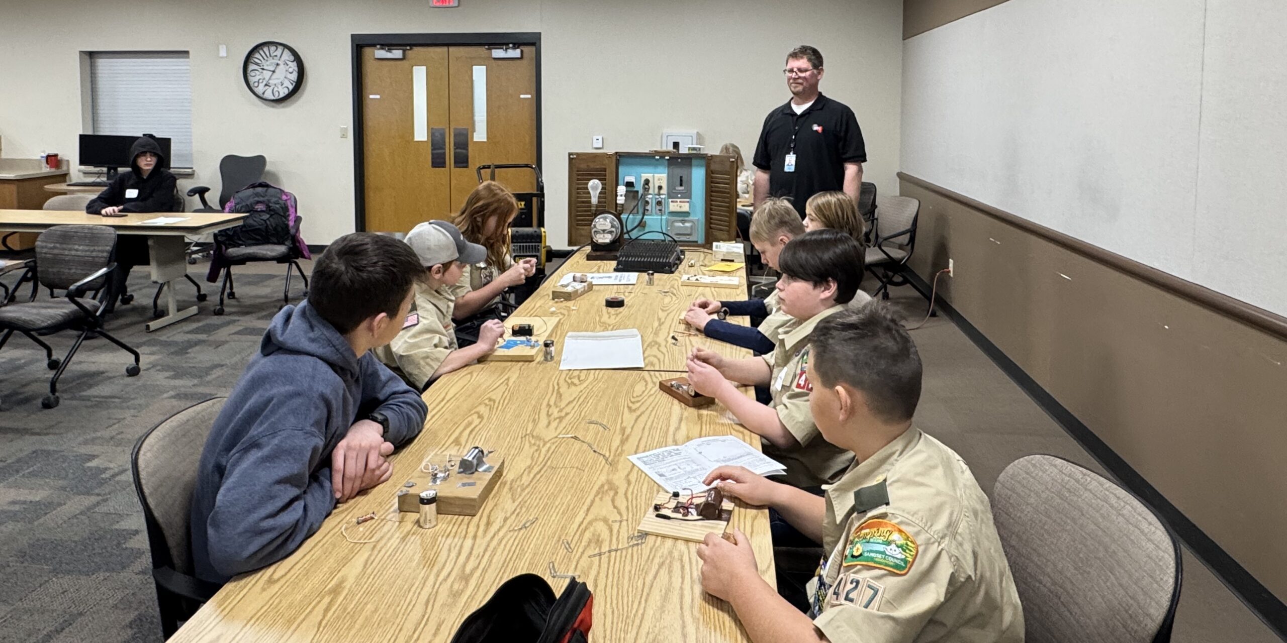 A WPS employee watches as a small group of Scouts test small electric circuits while sitting at a long table.