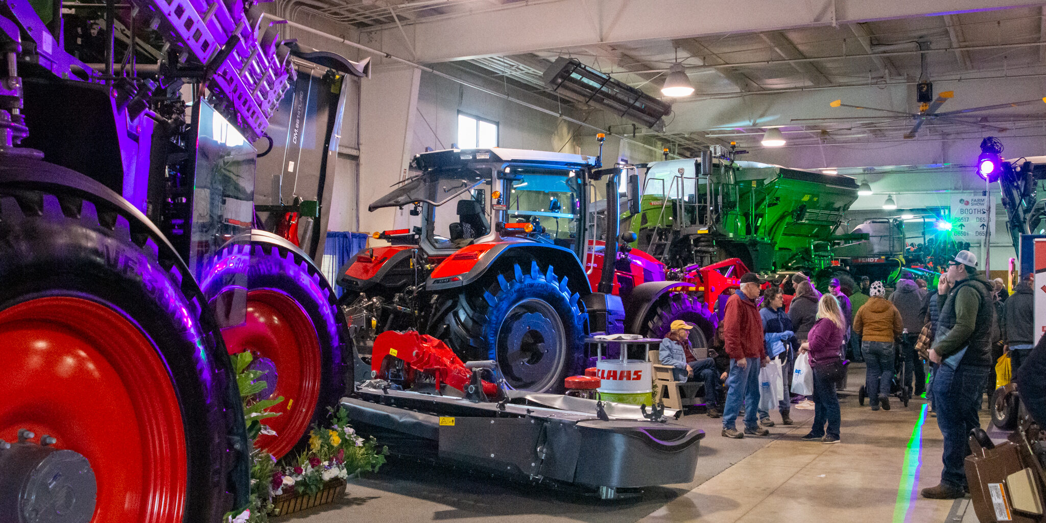 A small crowd of people look at and walk past several pieces of large farm equipment inside a warehouse-style building.