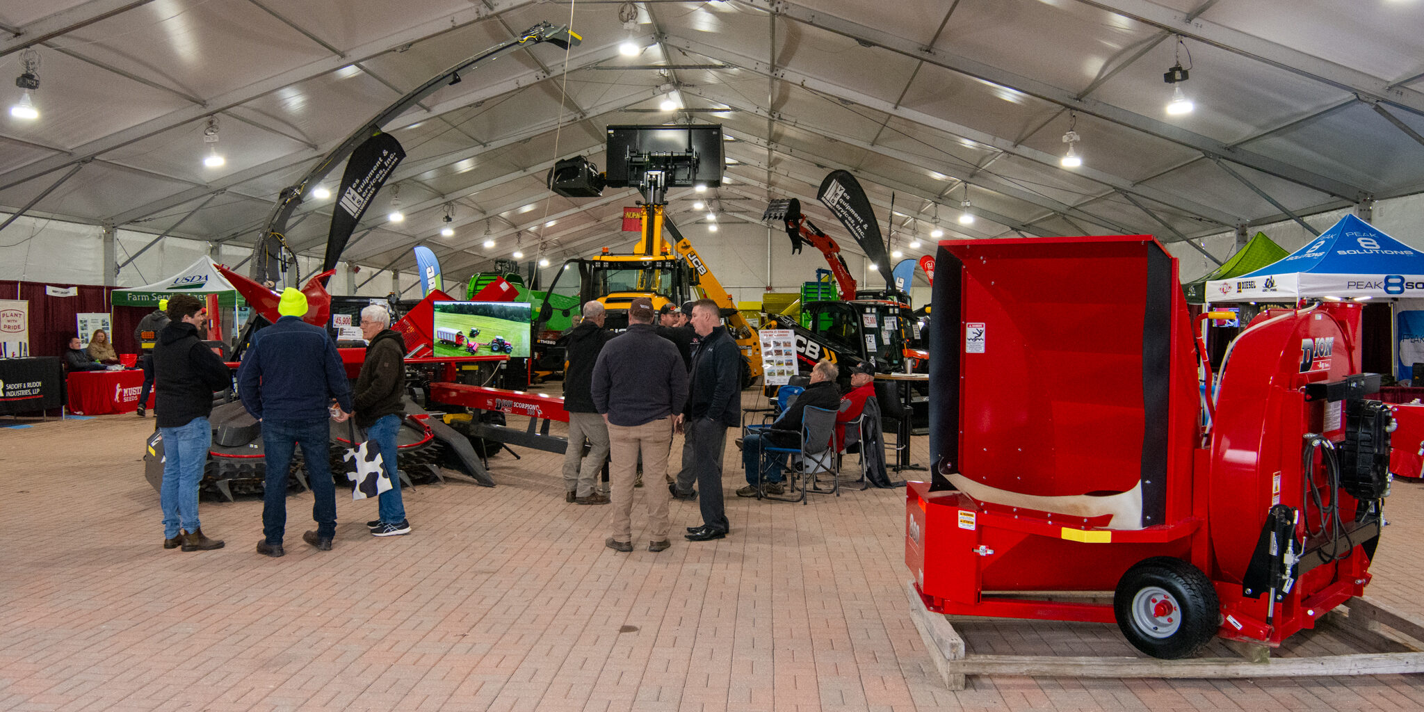 People stand near several pieces of large farm equipment inside a large exhibition tent.