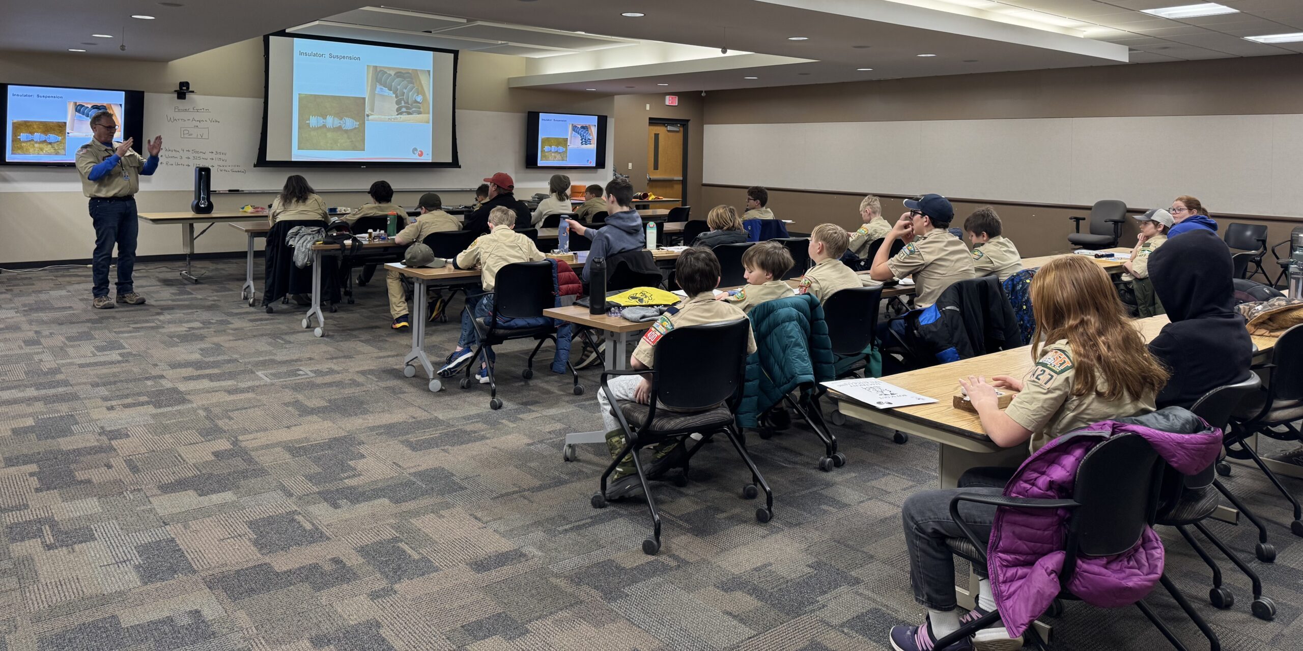 A group of Scouts sit at long tables listening to a WPS employee discuss electrical equipment in a large conference room.