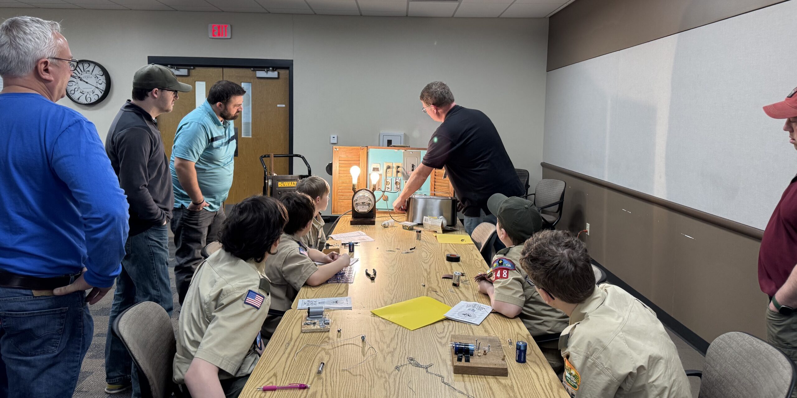 A group of WPS employees at the end of a long table show Scouts how electricity powers different household devices arranged on a small display board.
