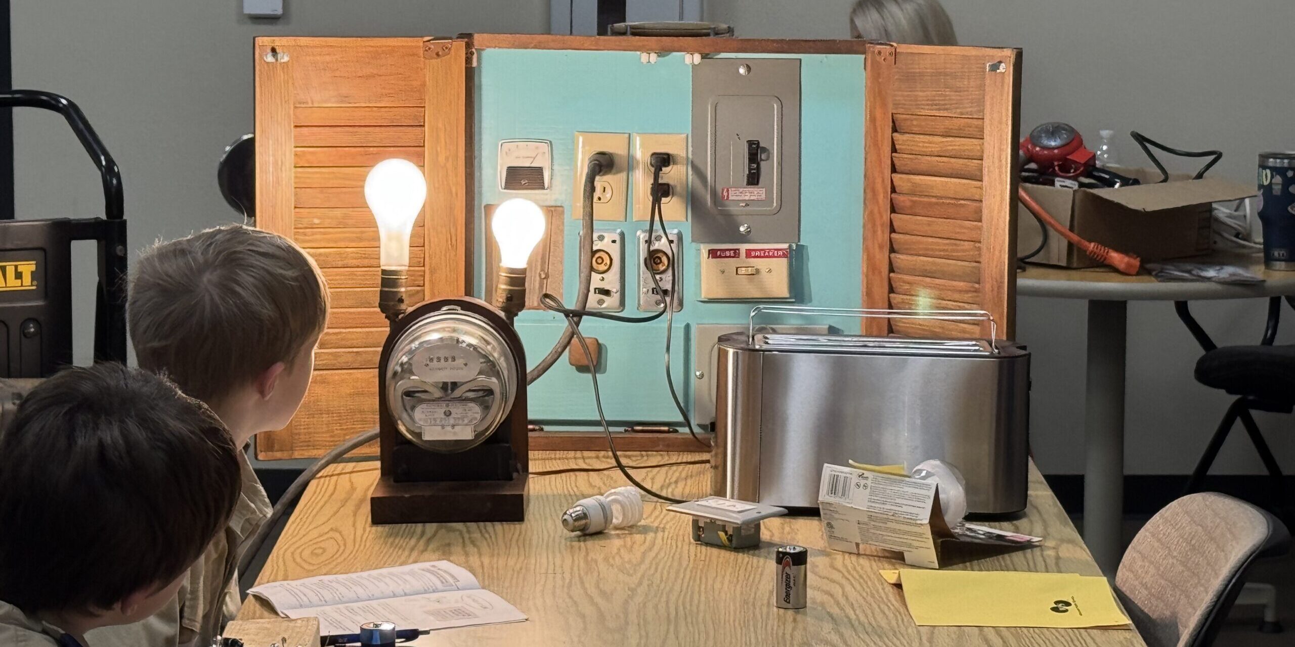 A pair of Scouts looks at an home electrical display featuring an electric meter, illuminated lightbulbs, a toaster, outlets and a circuit breaker.