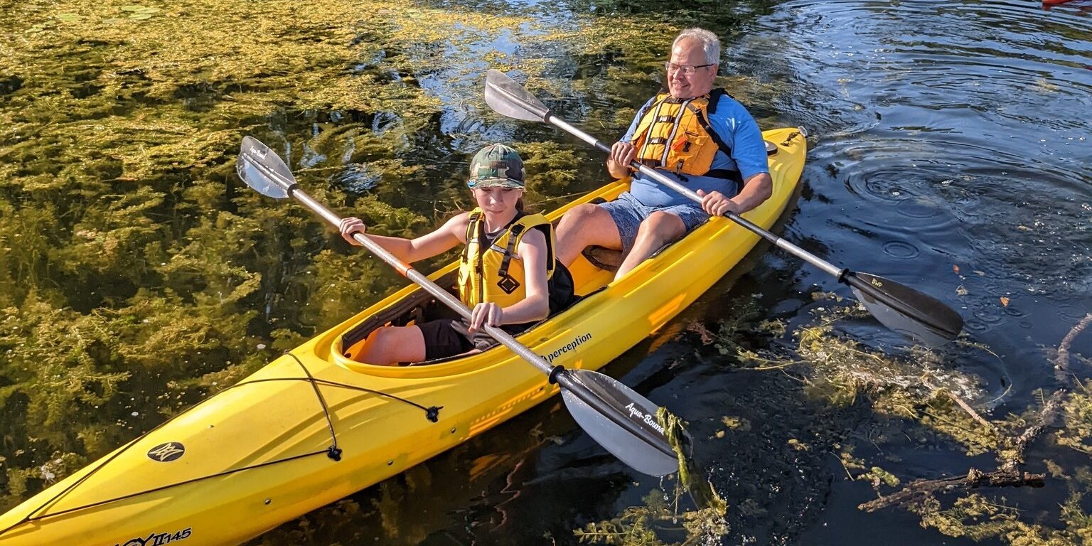 A man and a teenager paddle a kayak in a small pond on a sunny day.